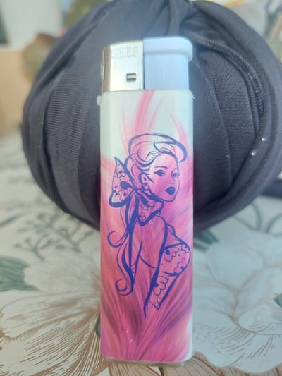 A close-up, eye-level shot shows a pink and white lighter with a drawing of a woman on it, positioned in front of a dark grey backpack. The lighter is upright, and the backpack is slightly out of focus behind it. The lighter has a silver top with "SEES" etched into it. The woman on the lighter is drawn in blue outline and features stylized hair, a bow, and clothing details. The background appears to be a table with a floral-patterned tablecloth. The lighting suggests an indoor setting, possibly daytime. The provided context places this scene in Markovo, Bulgaria.