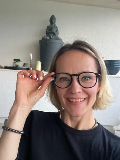 A woman with blonde hair, wearing black-rimmed glasses and a black top, is smiling and holding her glasses with her right hand. She has a beaded bracelet on her right wrist and a pearl necklace around her neck. In the background, to the left, is a gray-toned statue on top of a gray object with what appears to be a white candle, a corkscrew, and an item that is likely a plant. The background appears to be a plain, light-colored wall. The environment doesn't reveal information that would provide the location of Trebesin, Montenegro.