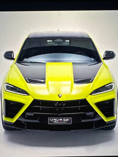 The image displays the front view of a bright yellow Lamborghini Urus SE parked on a white-ish surface, likely a showroom or studio. The vehicle has a black hood with a prominent yellow stripe running down the center, and a black grille with angular, aggressive styling. The headlights are sleek and modern, with distinct LED daytime running lights. A small rectangular badge on the lower grille reads "URUS SE Performance". The background is a plain, light-colored wall, suggesting a controlled environment rather than an outdoor setting in Sant'Angelo di Piove di Sacco, Italy. There are no people or other objects visible in the scene.