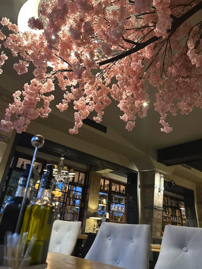 The image depicts the interior of a restaurant in Razlog, Bulgaria, characterized by an elegant and atmospheric setting. Dominating the upper portion of the frame are branches adorned with numerous artificial pink cherry blossoms, creating a visually striking canopy that hangs from the ceiling. The lighting is subdued, contributing to a cozy ambiance.
In the foreground, a table is set with what appears to be two bottles of olive oil and possibly balsamic vinegar, along with some utensils in a holder. The bottles are clear glass, with one containing a greenish liquid and the other a darker liquid, likely olive oil and vinegar.
Beyond the table, the restaurant's interior is visible. Several plush, light-colored chairs are arranged around tables, suggesting a dining area. In the background, large windows or glass partitions reveal shelves stocked with bottles and glassware, indicative of a bar or wine display. A chandelier with multiple lamps hangs in the mid-ground, providing soft illumination. The walls feature a combination of stone and darker paneling, adding to the restaurant's sophisticated decor. The overall impression is one of a well-appointed dining establishment, possibly aiming for a romantic or tranquil dining experience, enhanced by the floral overhead display.