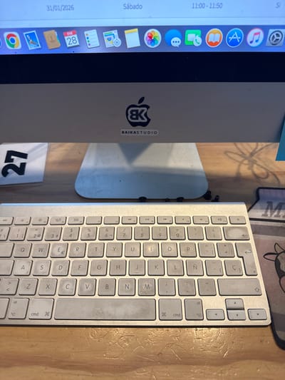 The image depicts an indoor scene featuring a desktop computer setup on a light-colored wooden desk. The central subject is an Apple iMac computer, with its silver-colored lower bezel and stand visible. Affixed to the lower bezel is a black sticker featuring a stylized "BK" logo within an Apple silhouette, and a smaller white rectangular sticker beneath it that reads "BAIKASTUDIO".
The iMac's display shows a macOS desktop environment. The top menu bar indicates the date "31/01/2026", the day "Sábado" (Saturday), and the time "11:00 - 11:50". A dock of application icons is visible, including Chrome, Mail (with a "28" notification on the calendar icon, possibly for ENE 28), Notes, Photos, Safari, and other standard macOS applications.
In front of the iMac, an Apple Magic Keyboard is positioned on the desk. The keyboard exhibits visible wear and dirt accumulation, particularly on the spacebar and several alphanumeric keys. The keyboard layout includes Spanish characters such as 'Ñ' and 'Ç'. Directly below the iMac's stand, several small, dark, cylindrical objects, possibly screws or debris, are scattered on the desk surface. To the left of the iMac stand, a white card or piece of paper with the number "27" printed in black is partially visible. To the right of the keyboard, a segment of a mousepad with a cartoonish cow graphic and the partial letter "M" is visible.
Based on the "BAIKASTUDIO" identifier and the Spanish keyboard layout, the location of this scene is identified as an office or studio in Puebla City, Mexico.