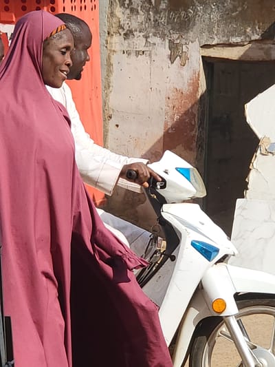 Two individuals are captured in an outdoor scene, likely resting in Maiburiji, Nigeria, under bright midday or early afternoon sun. In the foreground, an adult female lies on her back, covered by a deep maroon fabric that acts as both a body wrap and a head covering, similar to a hijab. A patterned orange and black band is visible around her forehead beneath the fabric. Her eyes are open, looking upwards, and her mouth is slightly open, suggesting consciousness but possibly weariness.
Behind her, an adult male, with dark skin and a close-cropped haircut, is also resting. He wears a white long-sleeved garment, and his head is visible next to the female's. Both individuals are lying on an orange, perforated plastic surface, which could be a mat, bench, or part of a stretcher. The male's right hand extends forward, grasping the handle or throttle of a white scooter that occupies the upper right portion of the frame. The scooter features prominent blue accents on its headlight and an orange circular reflector near its front wheel.
The background consists of a weathered building facade with light-colored paint peeling away to reveal a darker, possibly reddish-brown, layer beneath. The bright, high sun indicates clear weather and casts minimal shadows, reinforcing the impression of a daytime setting. The overall scene suggests a moment of pause or rest, perhaps during a journey or a break from activity, potentially due to the heat. No discernible text is visible in the image.