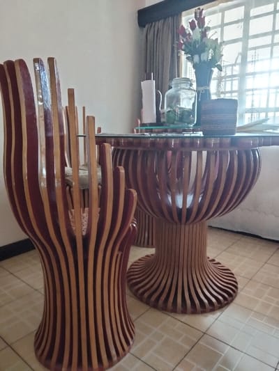 The scene is an interior room, identified as being in Riabai ward, Kenya, featuring a dining table and chairs. The floor is covered with light-toned square tiles. Two chairs and a round dining table are visible, all constructed with a distinct design of vertical, curved wooden slats that alternate between dark reddish-brown and lighter natural wood. The chairs have high, fan-like backrests and solid, flared bases. A light-colored cushion rests on one chair. The table features a circular glass top. Items arranged on the table include a white roll of paper towels on a brown stand, a large clear glass jar with a metal clasp lid containing green objects, a dark vase holding artificial red roses and white flowers, and a small, cylindrical woven container with horizontal stripes. A window with a visible grid pattern, partially covered by dark grey curtains and a valance, is present on the right, allowing bright natural light into the room. The walls are light-colored. No individuals are present, and no actions are depicted.