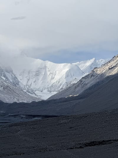 The image presents a view of a snow-covered mountain, partially obscured by clouds on the left, and steep, dark rock formations on the right. The photograph appears to be taken in a mountainous region, possibly within the vicinity of Tashi Dzom town, China, given the location context. The mountain peak is sharp and covered in snow, contrasting with the rugged terrain beside it. The cloudy sky suggests a high-altitude environment.