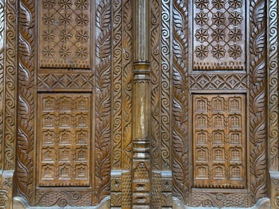 This image presents a detailed, frontal view of an intricately carved wooden surface located in Ghandruk, Nepal. The structure, likely a door or a large architectural panel, exhibits symmetrical relief carvings across its dark brown surface.
The central feature is a prominent vertical column-like element, which is narrower at its upper and lower sections, featuring a distinct capital and base adorned with vertical fluting and geometric patterns, including a section of small, raised, pointed shapes near its bottom. Flanking this central column are four narrower vertical panels, two on each side, uniformly carved with continuous, intertwining leaf and scroll motifs ascending vertically.
Two large rectangular panels are positioned in the upper sections, one on the left and one on the right, each displaying a grid of nine identical, eight-petaled floral motifs. Below these, intermediate horizontal bands feature repeating diamond-shaped patterns with internal crosses. Further down, two larger rectangular panels exhibit a grid of square or rectangular elements, each framed by an arched or trefoil-like surround. The lowest visible sections consist of horizontal carved bands with flowing scrollwork, wave-like textures, and a series of small, raised, elongated shapes.
The wood displays varying brown tones with visible grain and minor signs of wear, consistent with a polished or varnished finish. No people, objects other than the carved wood, or actions are depicted. The scene is a close-up, isolating the carved surface from its broader architectural or natural environment.