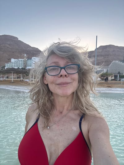 The media file shows a woman posing for a selfie while in the Dead Sea, in Tamar Regional Council, Israel. She has blonde, wavy hair and is wearing glasses and a red bikini. She is smiling gently at the camera. The Dead Sea is in the foreground, with its distinctive calm, bright green water. Behind her, there is a sandy beach with some structures and palm trees, and further in the background, there are hotels and brown, rocky mountains. The sky is a pale blue, suggesting it is either early morning or late afternoon. The overall mood appears relaxed and serene. There is no visible text in the image.
