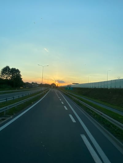 The image depicts a highway in Podłęże, Poland, during sunset. The camera angle is from a moving vehicle, looking forward and slightly to the right. The highway features multiple lanes separated by white markings. A metal guardrail runs along the right side of the road, bordered by a grassy embankment. A sound barrier, composed of vertical panels, extends along the left side of the highway, obscuring the view of any distant landscape.
The sky displays a gradient of colors, transitioning from a soft blue to warm oranges and yellows near the horizon, indicating the setting sun. A few streetlights are visible along the highway. In the distance, some trees and buildings are faintly visible beneath the sunset. The overall atmosphere suggests a peaceful journey as the day concludes.
There are no people visible in the image. No discernible text is present, though a green road sign with white lettering is partially visible in the distance. The weather appears clear, with the sunset creating a warm and serene ambiance. The scene is typical of a modern highway, with its orderly lanes and infrastructure.