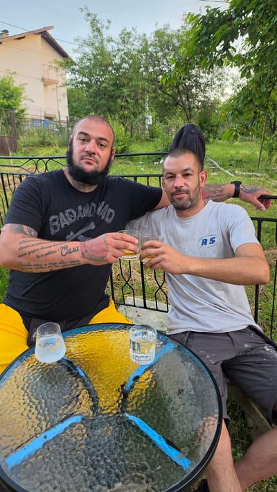 The image depicts two adult males seated outdoors at a small table, engaged in a toast, in what appears to be a residential area in Pernik, Bulgaria.
The man on the left is heavy-set, with a bald head and a full dark beard. He wears a black t-shirt with the text "BAD AND MAD" and crossbones, and yellow shorts. His arms are heavily tattooed with script and a prominent red rose on his left hand. He holds a clear glass containing an amber liquid in his right hand, extended towards the other man's glass. His left arm is draped over the shoulder of the man on the right.
The man on the right is of a slender build, with short dark hair and light facial stubble. He has a small red mark on the bridge of his nose. He wears a light grey t-shirt with a blue "RS" logo and smaller text, and dark grey shorts, possibly stained with paint. A black wristwatch is visible on his left wrist. He holds a clear glass with amber liquid in his left hand, clinking it with the glass of the man on the left. His right arm is draped over the shoulder of the man on the left.
Between the men, on a round, textured glass table, are two additional clear glasses; one is frosted and contains a clear liquid, and the other is clear and also contains a clear liquid. The table's underside features a design with blue and yellow/gold sections.
The background consists of a dark metal fence, lush green trees, and general vegetation. In the upper left background, a light-colored, two-story house with a brown roof is visible. The sky is light, indicating daylight conditions, likely late afternoon or early evening.