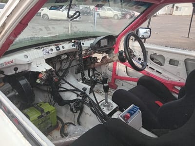 The image displays the interior of a heavily modified, stripped-down white vehicle, identified as a rally or race car, viewed from the passenger side looking forward through the windshield. A prominent red multi-point roll cage is installed, extending along the A-pillar and roofline. The original dashboard has been removed, revealing extensive exposed wiring, multiple electrical relays/fuse boxes, and a centrally mounted small digital display unit. An aftermarket black steering wheel with a quick-release hub is present on the driver's side. A single black racing bucket seat with red harness slots is installed on the passenger side. A tall, aftermarket gear shifter and an additional lever (consistent with a hydraulic handbrake) are mounted centrally on the floor. A green "AMARON" automotive battery is visibly secured in the passenger footwell, adjacent to a small white control panel equipped with blue and red rocker switches. The interior door panels are stripped, exposing internal metalwork. Through the windshield, an unpaved outdoor staging or service area is visible, containing several parked vehicles, including a white SUV, and distant low-rise buildings under daylight conditions. No individuals are depicted. The location is an unpaved outdoor staging or service area, likely within or on the outskirts of the city of Kisumu, Kenya.