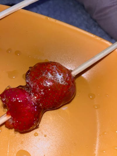 Two strawberries, coated in a translucent, hardened sugar glaze, are affixed to a single wooden skewer. The item rests on an orange, planar surface, likely a plate, located in Zlatovrah, Bulgaria. The top strawberry exhibits an irregular white, opaque crystalline substance on its upper surface. The lower strawberry displays a partially consumed or fractured area. Small, viscous liquid droplets are distributed across the orange surface. A blurred, dark-colored textile or soft material occupies the background. No individuals are present.