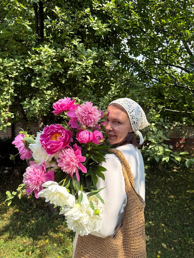 A woman with dark hair, wearing a white crocheted headscarf and a cream-colored sweatshirt, stands outdoors holding a large bouquet of pink and white peonies. She is looking towards the camera and smiling. The bouquet is positioned in front of her, obscuring part of her body. She is carrying a woven, tan-colored bag over her right shoulder. The background consists of lush green foliage from trees and a hint of a brick structure on the right. The ground is covered in green grass with scattered fallen yellow leaves. The scene appears to be a garden or yard, likely in a rural or suburban setting, possibly within the Прикамский district of Russia, given the context.
