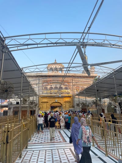 The image depicts a view of the Golden Temple, also known as Sri Harmandir Sahib, in Amritsar, India. A large number of people are walking on a pathway lined with golden railings. The pathway leads towards the main entrance of the temple, which is adorned with intricate gold work. Above the entrance, text in a language other than English is visible. The temple itself has a dome and detailed architectural features. Overhead, a metallic structure with fans and a canopy extends over the pathway, providing shade. The sky is clear and blue. People are dressed in various attire, with many wearing turbans, indicating a religious or cultural gathering. Some individuals are carrying bags. The overall scene suggests a busy day at a significant religious site.