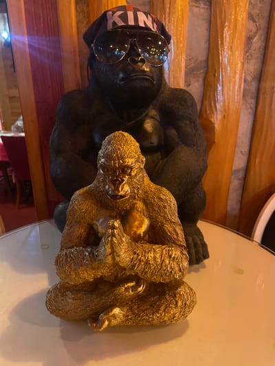 The image displays two gorilla statues positioned on a reflective white tabletop in an indoor location within Moračko Trebaljevo, Montenegro. In the foreground, a smaller, golden-bronze colored gorilla statue sits with its legs crossed and hands pressed together in a meditative pose. Its surface exhibits a textured finish. Behind it, a larger, dark-colored gorilla statue is seated, adorned with aviator-style sunglasses and a bandana featuring the word "KING" in white lettering on its forehead. The background consists of vertical wooden panels or log-like structures with a warm, light brown hue. Reflections of interior lighting and indistinct red furniture are visible on the tabletop and in the sunglasses.