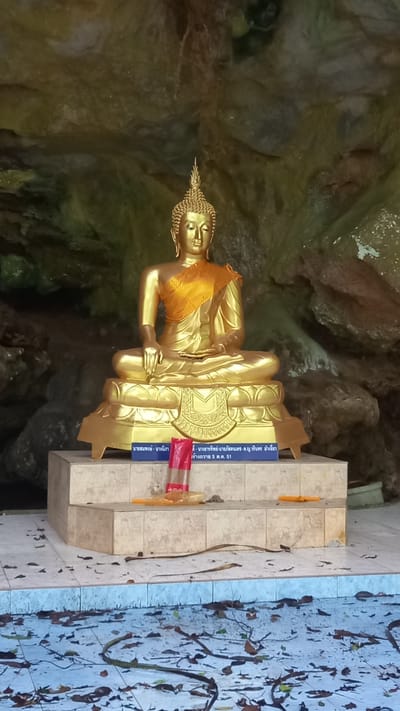 A gilded statue of Buddha sits in meditation on a multi-tiered beige tile base. The statue wears a yellow cloth draped over its shoulder. A yellow candle is placed on the step to the statue's right. Red-and-white striped tape and signage written in Thai are on the step in front of the statue. The statue is placed against a backdrop of mottled rock formations. Dead leaves litter the concrete floor. The city of Ban Pak Ra is not visible in the image.