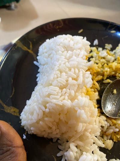 On a black plate, two kinds of rice are visible, white rice molded into a rectangle and yellow rice scattered next to it. A metal spoon rests on the yellow rice. A dark-skinned hand is visible in the lower right corner of the image. The image does not contain any information about the city of Nsukka, Nigeria.
