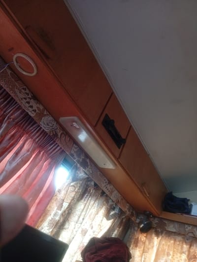 The image captures an interior view, likely within a recreational vehicle or a mobile home, displaying a section of cabinetry and curtains. The cabinetry is constructed of wood with a light brown finish, and it runs along the top of the frame. On the left side of the cabinetry, a circular silver object, possibly a hook or a decorative element, is visible. Below the cabinetry, patterned curtains are drawn, with a bright light source emanating from the right side, suggesting a window or an opening. A long, rectangular light fixture is mounted horizontally on the cabinetry, and to its right, a black vent or grille is embedded in the wood. On the far right of the frame, a dark blue folded fabric item, possibly clothing or a bag, rests on a shelf. The overall impression is of a lived-in, functional space. The location of West Suffolk, United Kingdom is not discernible from the visual elements present in the image.