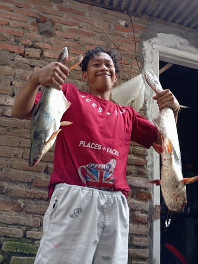 The image shows a man standing in what appears to be an outdoor or semi-outdoor setting, possibly in the city of Baron, Indonesia, judging by the appearance of the surroundings. He is holding two large fish, one in each hand. The man is wearing a red t-shirt with white lettering and a graphic, and light-colored pants. The background consists of a brick wall and a partially visible window or doorway. There's also a white plastic bag visible behind the man. The lighting suggests it is daytime.