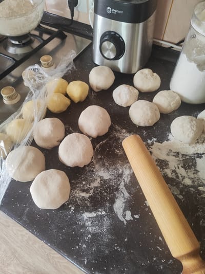 The image depicts a kitchen scene with several balls of dough arranged on a black countertop. A wooden rolling pin lies diagonally across the lower right portion of the frame. In the background, a stainless steel blender is visible, along with a glass jar filled with white powder, presumably flour. To the left, a glass bowl containing more dough is on a stovetop. The overall environment suggests food preparation. The location in Gvardeysk, Russia, is not visually determinable from the image itself.