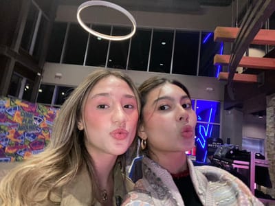 Two women pose indoors, their faces angled to the camera in a selfie-style composition. One wears a patterned shirt, while the other is in a tan garment. Both have fair skin and styled hair, pursing their lips slightly. In the background, there's a multi-level interior with windows, lighting fixtures, and branding elements. The entire scene is rotated, giving an unusual perspective.