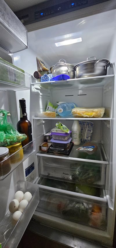 The image shows the interior of a refrigerator, viewed from above. The refrigerator is organized into several compartments. On the left, there are two large metal pots stacked on top of each other. Next to them, there are some food items wrapped in plastic, including what appears to be cheese. Further to the right, there is a large blue water jug, and next to it, more food items in plastic packaging and containers. One container has purple lid and is filled with green leafy vegetables. In the middle section, there is a brown bottle, possibly containing a beverage, lying horizontally. Next to it, a green plastic bag is visible, along with other food items in clear containers. On the far right, there is a clear plastic drawer containing several white eggs. The refrigerator is illuminated by internal lights, which are on. There is no visible indication of the time of day, weather, or emotions. The location is stated as Vinnytsia, Ukraine. Some visible text on the containers and packaging is not legible due to the image quality and resolution.