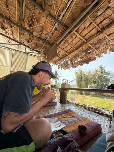This image depicts a male seated within a rustic, open-air, elevated establishment in Pai, Thailand. The man, with dark curly hair, a mustache, and a short beard, wears a two-tone baseball cap (dark purple/grey and black), a dark short-sleeved t-shirt, and dark shorts with a green stripe. Tattoos are present on his right forearm. He is actively consuming a light-colored sandwich, holding it with both hands. He is seated cross-legged on a multi-colored patchwork cushion placed on a woven mat floor. A clear plastic water bottle rests on a low, dark wooden table in front of him. A purple backpack is positioned near his feet. An ashtray containing cigarette butts and a white paper/napkin with crumbs are on the cushion to his right. The structure features a roof constructed from dried leaves and bamboo, supported by metal beams. A wall-mounted fan is visible on the left. The exterior view, beyond a wooden railing, shows a verdant field, palm trees, bamboo stalks, and distant hills beneath a clear, bright sky. A wooden condiment caddy is placed on the railing.