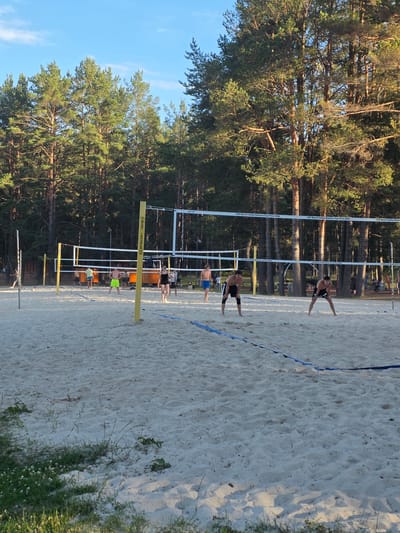 This is a landscape-oriented image of a beach volleyball court. The scene is outdoors, set against a backdrop of tall pine trees and a clear blue sky, suggesting a warm, sunny day, likely late afternoon given the long shadows. The sand on the court is light-colored and appears well-maintained, with distinct footprints visible.

The main subjects are several people playing or spectating a beach volleyball game. Two male athletes are actively engaged on the court, dressed in athletic shorts. One is in motion near the net, mid-play, while another stands further back. Other individuals are visible in the background, some appearing to be players, others perhaps spectators or staff, though their distance and the angle make precise identification difficult. The volleyball net, a key object, is clearly visible, dividing the court. Yellow poles with black lettering mark the sides of the court.

The text "CEV Beach Volley Nations Cup Final 2024" is visible on the net poles, indicating the specific event taking place. Another piece of text, "SPORT TRANSFER," is also visible on a yellow support pole. The presence of the volleyball net, the players, and the court markings confirm the activity is a beach volleyball match. The overall atmosphere appears to be energetic and competitive, typical of a sports event. The setting in Daugavpils, Latvia, provides the geographical context for this beach volleyball tournament.