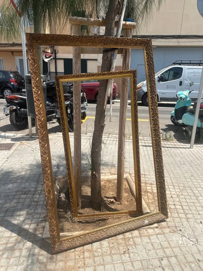 In Palma, Spain, two large, ornate golden picture frames are propped up against a tree on a tiled sidewalk. The frames are empty, and their intricate patterns are clearly visible. Behind the frames, a dark-colored car and a scooter are parked on the street. Further in the background, a white van with "CARPINTERIA" written on its side and a light blue scooter are parked. The scene is set on a sunny day, with shadows cast on the pavement.
