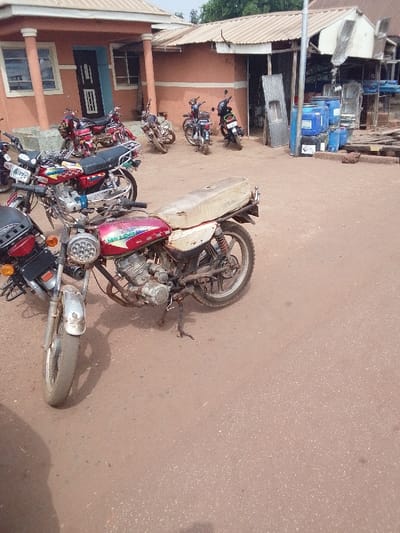This image captures an outdoor street or lot scene in Ankpa, Nigeria. The foreground is dominated by a weathered red and white "DAYLONG" brand motorcycle, identified by text on its fuel tank, parked on its kickstand on a reddish-brown unpaved surface. This motorcycle exhibits significant wear, a faded paint job, rust on metallic components, a torn cream-colored seat, and a multi-LED headlight. A portion of another black motorcycle is visible to the left. In the midground, approximately five to seven additional motorcycles of various models are parked in an unorganized manner. The background features two single-story buildings. The structure on the left has a light brown/orange painted facade, white-framed windows, and a dark blue or black door with white paneling, topped by a light-colored corrugated metal roof with an overhang. Adjoining it on the right is another single-story building with a similar facade and corrugated metal roof, appearing to be an open-fronted shop or workshop. A utility pole stands near this second structure, adjacent to which are stacked blue plastic barrels (at least five visible), metallic sheets, and wooden planks. The sky is overcast. No individuals are present in the scene.