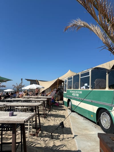 This image captures a sunny day at a beachside establishment in Zandvoort, Netherlands. In the foreground, a row of empty wooden tables and chairs on a sandy beach are bathed in sunlight, casting long shadows. To the right, a vintage green and cream bus, identified with the text "WESERBERGLAND-KLINIK HÖXTER," is parked on a concrete ramp. The bus appears to be part of the establishment, possibly serving as a bar or dining area.
In the background, a large, beige, tent-like structure offers shade over a seating area with white umbrellas and numerous people. A few palm trees, one dry and sparse, add to the beachy atmosphere. The clear blue sky suggests fair weather. The scene depicts a relaxed outdoor dining or lounge area by the sea, with a unique bus as a central feature.