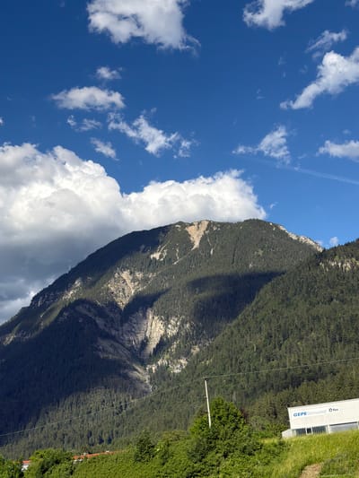 A wide-angle, eye-level shot captures a majestic mountain in Farchant, Germany, under a bright blue sky dotted with white cumulus clouds. The mountain, covered in a dense forest of dark green coniferous trees, rises steeply, with a lighter, rocky scar visible on its upper slope. A dramatic shadow casts a large portion of the mountain into darkness. In the foreground, a small, modern white building with the blue text "GEPE Geimupast Q flex" on its side is situated on a grassy incline. A thin utility wire stretches horizontally across the frame in front of the mountain. The overall scene depicts a natural landscape with a touch of industrial presence.