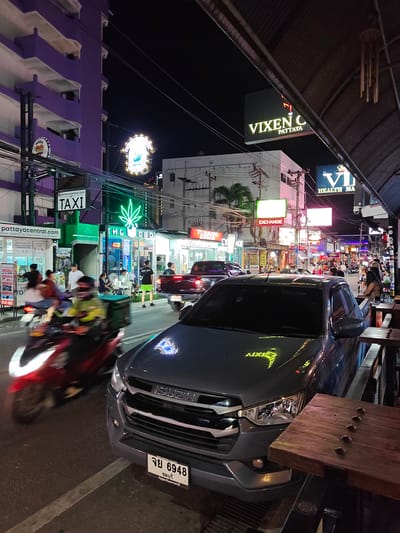 This is a nighttime street scene in Pattaya, Thailand, characterized by bright neon signs and illuminated buildings. The foreground features a dark grey Isuzu pickup truck, with its license plate displaying Thai script and the number "6948". To the left of the truck, a red motorcycle is captured in motion, with its rider partially visible. The street is lined with shops and businesses, including a "TAXI" stand and a store with a prominent green neon cannabis leaf logo, displaying "THC CBD". Signs for "VIXEN C PATTAYA", "EXCHANGE", and "HEALTH MA" are visible further down the street, indicating a lively commercial and entertainment district. People are seen walking and sitting at outdoor tables, suggesting a vibrant evening atmosphere. The overall mood is energetic and bustling, typical of a tourist area at night.