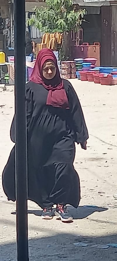 The image shows a woman standing in what appears to be a street or market setting, likely in Khan Yunis, Palestinian Territories. She is the central subject, dressed in a black abaya and a deep red or maroon hijab that frames her face. To her right, a small cart or trolley laden with various packaged goods is visible.
In the background, to the woman's left, there are stacks of light brown plastic chairs and colorful plastic items, possibly containers or other household goods, arranged on what look like market stalls. Green foliage from a bush or tree is also present. Further back, parts of buildings can be seen. A dark, horizontal bar crosses the lower foreground of the image, suggesting the photo was taken from an elevated position or through a barrier like a railing or window frame.
The scene takes place outdoors during the daytime, with clear lighting indicating sunny weather. The ground is an unpaved surface or worn concrete. The overall atmosphere suggests a bustling commercial area or a street market. The woman's expression is neutral, and she appears to be looking generally forward or slightly towards the camera. No legible text is visible anywhere in the image.