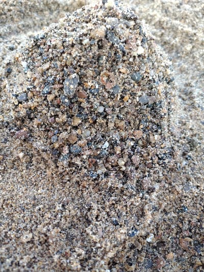 The image shows a close-up, overhead view of a pile of wet sand with visible pebbles of various sizes and colors. The sand is a mixture of light brown and grey grains, with darker, multi-colored pebbles interspersed throughout. Some pebbles are dark grey or black, while others are lighter brown, tan, or even reddish. The texture appears granular and slightly clumpy due to the moisture. The background consists of more of the same sandy material, also appearing wet and creating a soft, blurred texture. There are no people, animals, or man-made objects visible in the image. The overall scene is a natural, granular surface. It is not possible to determine the specific location within Saltburn by the Sea from this image.