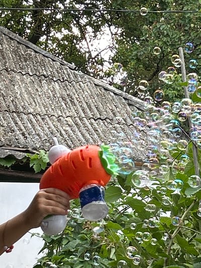 The image shows a hand holding a carrot-shaped bubble gun, which is expelling a stream of colorful bubbles into the air. The background consists of lush green foliage, a weathered gray roof with a corrugated texture, and a light-colored wall. The lighting suggests it is daytime. The scene appears to be outdoors, possibly in a residential backyard or park. There is no specific indication of the city of Prokhladny, Russia, or any other urban context within the visual information provided.