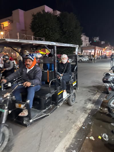 The image depicts a nighttime urban street scene in Jaipur, India. A black e-rickshaw is prominently featured in the mid-ground, oriented as if moving towards the viewer's right.

The e-rickshaw carries two individuals. The male driver, positioned on the left, wears a dark puffer jacket, blue jeans, an orange and blue knit hat, and light-colored sandals. His hands are on the handlebars. To his right, a female passenger sits, dressed in a black long-sleeved shirt, black pants, and white sneakers. Her lower face and head are covered by a light-colored patterned scarf, and she wears glasses.

In the left background, another individual on a dark two-wheeler (motorcycle or scooter) with a helmet is visible behind the e-rickshaw. Behind them, a multi-story building with a distinctive illuminated arched window feature stands against the night sky, partially obscured by dark green trees.

The right side of the image reveals a street extending into the distance, lined with buildings illuminated by various artificial light sources, suggesting commercial activity. Other vehicles, including cars and additional two-wheelers, and pedestrians are present on this street. The paved ground in the immediate right foreground appears wet or reflective and contains scattered debris. A parked dark scooter or motorcycle is partially visible on the far right edge of the frame.