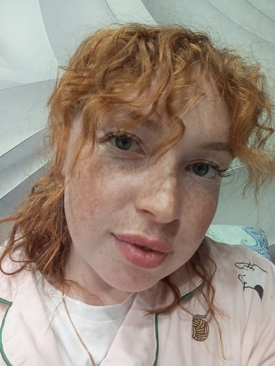 A close-up, slightly angled portrait shows a fair-skinned person with red curly hair and freckles. The person is wearing a light pink shirt with a white collar and a green trim. The shirt has cartoon-like designs on it, including a cat and a ball of yarn. A delicate gold chain necklace is visible. The background is a gradient of light gray and white. The location cannot be determined from the given image.