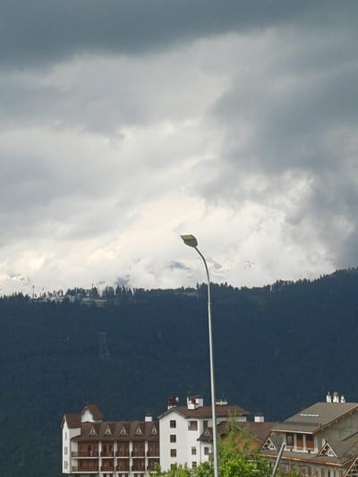 The image displays a cluster of buildings nestled on a densely forested mountainside, possibly a resort or village. The architecture suggests a European style, with brown tiled roofs and white or light-colored facades featuring numerous windows and balconies. Some buildings appear to be hotels or apartment complexes, with multiple stories.

In the foreground, a streetlamp with a curved arm is visible, pointing towards the sky. The sky is overcast with dramatic, cloudy formations, suggesting either early morning or late afternoon, or a generally cloudy day. The dense, dark green of the forest covers the steep slopes of the mountain, creating a stark contrast with the sky and buildings.

The orientation of the image is vertical, as if it was taken from a lower vantage point looking upwards, or simply rotated. The lighting suggests diffused daylight, with no strong shadows indicating direct sunlight. The overall mood is somewhat subdued due to the cloudy weather, but the presence of the buildings hints at human activity and habitation within the natural landscape. No people are clearly visible, and there is no discernible text in the image. The scene is set in a mountainous, likely rural or semi-rural, environment.