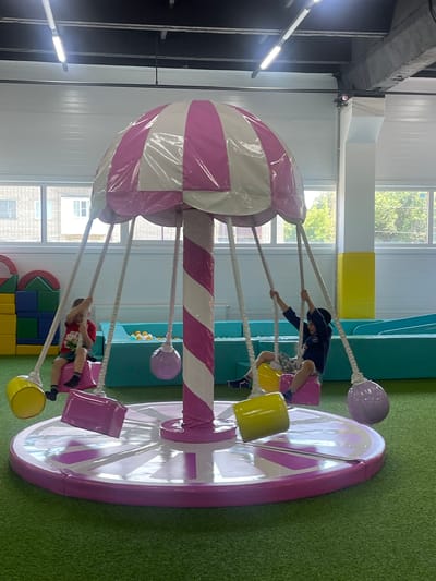 A brightly lit indoor playground features a central carousel with a pink and white striped umbrella top, resembling a candy. Two young boys are on the carousel. The boy on the left, wearing a red t-shirt and green shorts, sits in a pink swing, holding the ropes. The boy on the right, wearing a dark blue hooded jacket and patterned shorts, is also in a pink swing, with his arms raised. The carousel has a pink circular base with a white pattern of radiating lines. Several colorful, spherical weights hang from the carousel's structure, including yellow, pink, and purple. The ground is covered in artificial green turf. In the background, a ball pit with blue and turquoise walls is visible, along with large colorful building blocks on the left and a yellow block structure and a blue slide on the right. Windows in the background show trees and buildings, suggesting an urban setting. The scene appears to be located in a public recreational facility in the city of Donskoye, Russia.