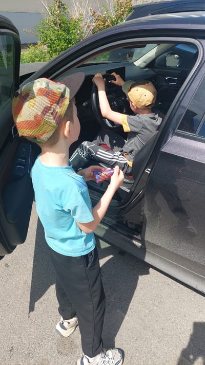 Two young boys, wearing baseball caps, are interacting with the interior of a parked car. The boy on the left, wearing a camouflage and orange cap, a light blue t-shirt, and black sweatpants, stands outside the open driver's side door, holding a small colorful cylindrical object in his right hand. The boy on the right, seated in the driver's seat and wearing a tan baseball cap, a grey t-shirt with orange accents on the sleeve, and black pants with white stripes, has his hands on the steering wheel, appearing to play. The car is dark colored and parked on an asphalt surface. In the background, there is green foliage and some dry brush. The scene suggests a casual outdoor setting, possibly a driveway or parking area. The specific location of Komsomolsk-on-Amur, Russia is not determinable from the visual information in the image.