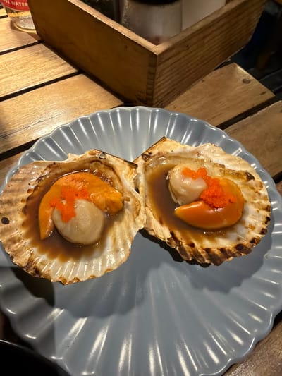 This media file is a close-up, overhead shot of two baked scallops in their shells, served on a grey, scalloped-edged plate. The scallops are cooked in a savory sauce, and each is topped with bright orange roe. The shells themselves are golden brown and appear slightly charred in places, suggesting they were baked or broiled.

The plate is resting on a wooden surface, likely a table, with planks of wood visible. In the background, more wooden elements are present, arranged in a way that suggests a rustic or outdoor setting. The lighting is warm and appears to be artificial, possibly from overhead restaurant lighting.

The scene captures a moment of enjoying a meal. There are no people visible. The focus is entirely on the food, emphasizing its appetizing presentation. The bright orange roe adds a pop of color and a sense of fine dining.

A partial view of a red and white label is visible on the bottom left, showing the text "0% WATER". This could indicate a beverage or a food product nearby, but its context is unclear.

Given the location context of Ko Tao Subdistrict, Thailand, this dish could be a local delicacy or a popular seafood offering in a restaurant there. The overall impression is one of a delicious and well-prepared dish, ready to be enjoyed.