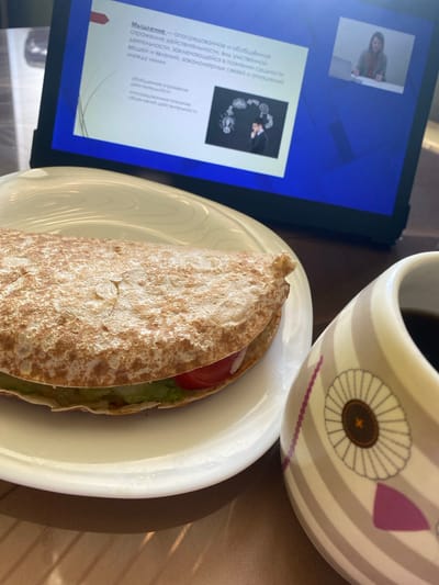 The image shows a close-up view of a partially eaten wrap on a white plate, next to a mug with a striped pattern and a floral design. In the background, a tablet or laptop displays a presentation with text and an image of a person. The presentation text appears to be in Russian and discusses "thinking" and "reflection." The overall scene suggests a casual meal or break while working or studying. The lighting is soft, indicating it could be daytime. The location is identified as Podolsk, Russia, but no specific environmental cues are visible in the image itself. There are no strong emotional cues, but the scene appears calm and focused on the meal and the device.
