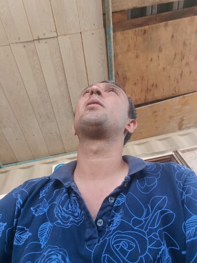 A low-angle, medium shot captures a man wearing a dark blue collared shirt with a light blue floral pattern, looking upwards. The man has short, dark hair with some graying and stubble on his face. The background consists of a wooden ceiling, possibly made of planks, and a section of unfinished wooden material. A rusty blue metal pipe is visible on the left side of the frame, and a glimpse of a white corrugated structure can be seen in the background on the right. The scene appears to be outdoors, possibly in an area under construction or a workshop, in Baku City, Azerbaijan.