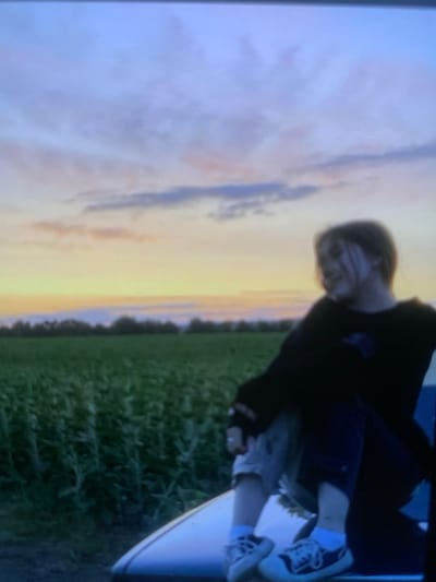 A young person with light brown hair, wearing a black sweatshirt, grey shorts, and white sneakers with blue accents, sits on the edge of a vehicle. They are looking to the right, with their body angled slightly away from the camera. The background features a field of green crops, a line of trees, and a sky filled with wispy clouds in shades of blue, pink, and orange, indicative of a sunset. The setting appears to be a rural area, possibly on the outskirts of Hoholeve, Ukraine.