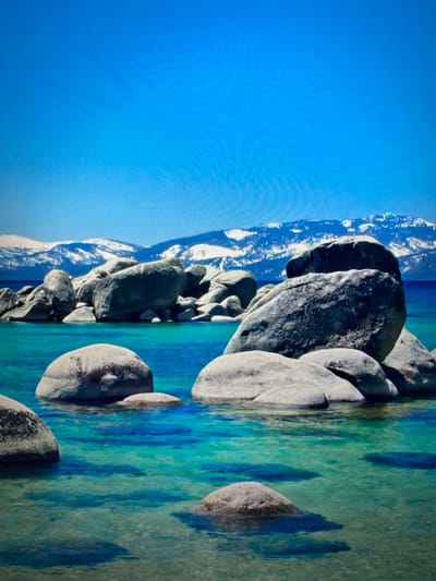 The image depicts a serene natural landscape. In the foreground, several large, smooth, grey boulders are partially submerged in clear, turquoise water. The water is calm, with gentle ripples and shadows visible beneath the surface, indicating shallow depths in some areas.
In the mid-ground, a cluster of larger, similarly shaped boulders forms a small rocky outcrop. These rocks are also grey and appear weathered. Behind these rocks, the water transitions to a deeper blue.
In the background, a mountain range with snow-capped peaks stretches across the horizon. The mountains are majestic and appear to be distant. The sky above is a clear, vibrant blue, devoid of clouds.
The overall scene is one of natural beauty and tranquility, characterized by the contrast between the rugged rocks, the pristine water, and the distant snow-covered mountains under a bright blue sky. The location is not in the city of Valencia, Spain, but rather appears to be a lake or coastal area with large rock formations.