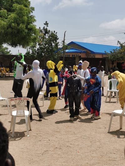 The image depicts a group of children and young adults playing a game in a schoolyard in Gerin Gada, Nigeria, located at the coordinates 9.93 latitude and 8.86 longitude.  There are multiple white plastic chairs in the background, with the writing "77W" visible on the top of one chair.  One person is wearing a black bodysuit, and another person is dressed in a yellow and black bee costume.  A third person is dressed in a Superman costume. The group of children are all gathered together playing a game. The surrounding environment is comprised of dirt and sand. There is a blue building in the background. The sky is clear and bright.  The photo was likely taken in a school setting. There are many other people in the background, and they are all smiling and looking happy.  The children are dressed in costumes for a performance.