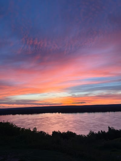 A wide shot captures a twilight sky over a tranquil body of water. The sky is a vibrant canvas of color, with hues of deep blue transitioning to soft pinks, oranges, and lighter blues as they approach the horizon. Wispy clouds are scattered across the sky, catching the last rays of the sun and adding texture to the scene. A dark, silhouetted treeline stretches across the middle ground, separating the sky from the water. The water itself reflects the colors of the sky, creating a shimmering, pastel surface. In the foreground, dark green foliage forms a dense embankment that slopes down towards the water's edge. This image appears to be a natural landscape, possibly a river or lake, and is not specifically identifiable as being located within the city of Novokuybyshevsk, Russia, based solely on visual information. There are no people, animals, or man-made structures present in the image.