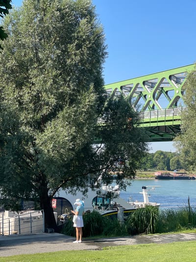 A full, sunny shot captures a woman standing near a large tree by a body of water, under a green truss bridge. The woman, seen from behind, wears a white skirt, a light blue top, and a light-colored hat with a wide brim. She holds a mobile device in her hands. To her left, a large tree with dense foliage dominates the foreground. Behind the tree and woman, a docked boat is visible, with the boat's name partially obscured but reading "PIR". The water is a bright blue, and across the water, a park-like area with trees lines the opposite bank. In the background, the distinctive green truss bridge, likely part of a railway line, spans the water. The sky is clear and blue. The overall scene suggests a leisurely day in Bratislava, Slovakia, potentially near the Danube River, with the bridge being the Apollo Bridge given its characteristic green color and truss design.