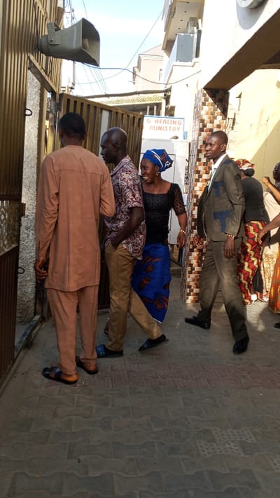 People, dressed in varied attire, walk and stand outside a building. A man in a brown suit is in mid-stride, a woman with a blue headwrap smiles, while two other men stand near a gate. A sign reads "& Healing Ministry." Overhead, a large speaker is mounted on the gate. The setting appears to be an outdoor street with buildings on either side.