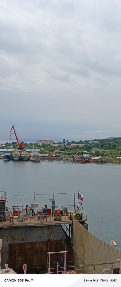 The image captures a waterfront scene in Nakhodka, Russia, under an overcast sky. In the foreground, a dilapidated ship or barge with rusty metal plating occupies the lower portion of the frame. The deck of this vessel is cluttered with various industrial equipment, including pipes, containers, and safety railings. A Russian flag is visible on the right side of the vessel's deck.

Beyond this foreground vessel, a calm body of water stretches towards a harbor. Several fishing trawlers or similar vessels are moored in the middle ground, displaying a worn and weathered appearance. A large red industrial crane stands prominently on the shore, indicating ongoing port activities.

In the background, a coastal town unfolds with a mix of residential buildings, churches with blue domes, and abundant green foliage. The overall atmosphere is one of a working port and a coastal settlement under a somber sky. The image appears to be a photograph taken from a slightly elevated viewpoint, looking down towards the water and the town.