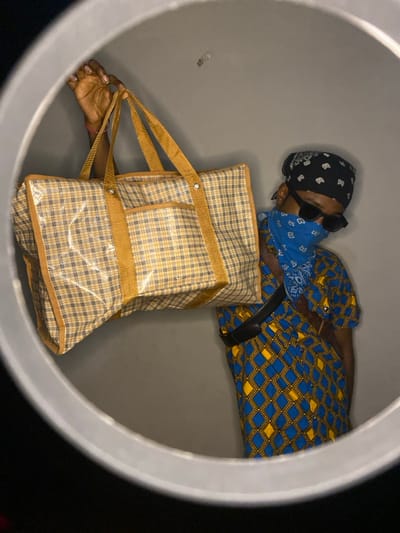 A person wearing sunglasses, a bandana, and a blue patterned mask, reclines inside a circular container, possibly a large tube or barrel. They wear a blue and yellow patterned shirt and a black belt. A large plaid shopping bag with transparent pockets is also inside the container. The person's hand holds the bag's straps. The inside of the container is gray, while the exterior is dark.